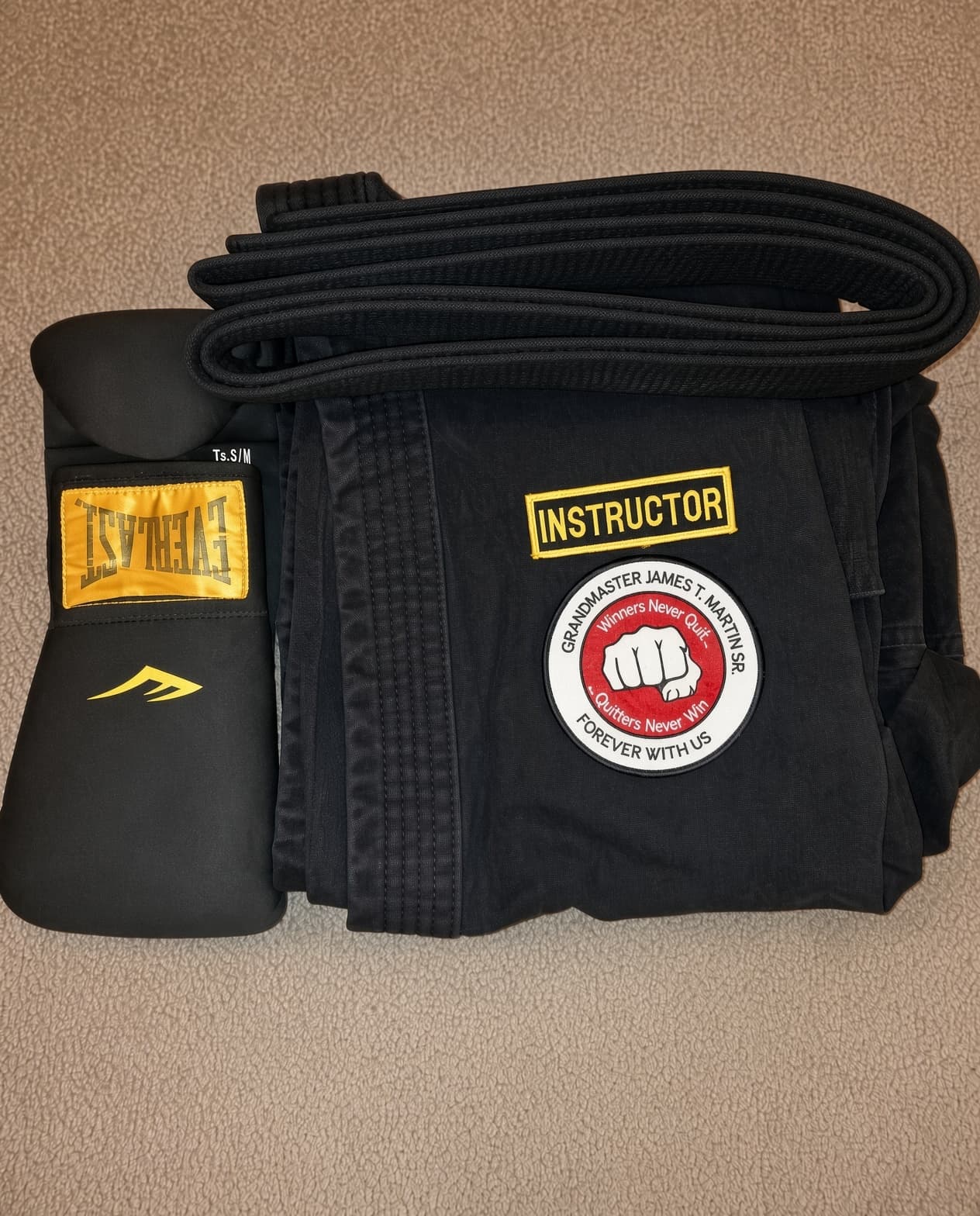 Photograph of folded martial arts gear: a black belt, a black uniform top carrying a yellow patch reading INSTRUCTOR and a circular patch reading Grandmaster James T. Martin Sr., Winners Never Quit, Quitters Never Win, Forever With Us, and a black Everlast glove.